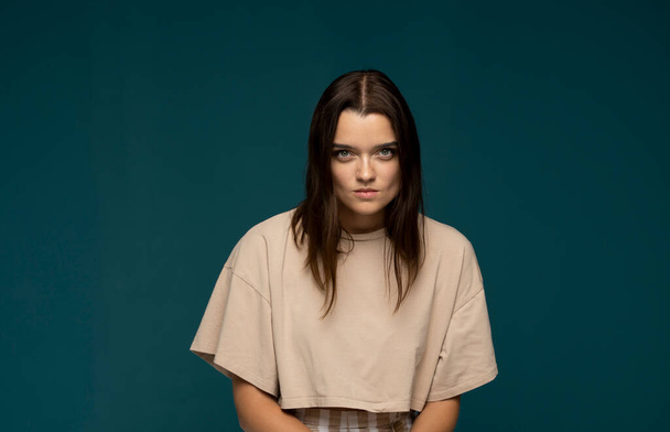 Portrait of an upset, angry young brunette girl in a beige oversize t-shirt looking in a camera isolated over blue background. Emotions. - Valokuva, kuva