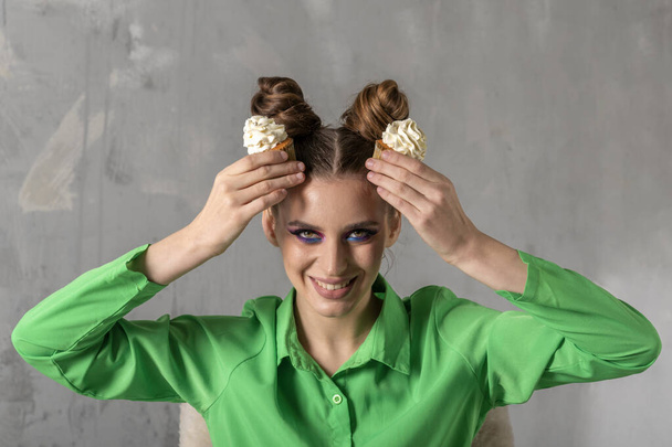 Smiling young woman with two buns hairstyle holds two cupcakes above her head. Portrait of girl in green blouse with mafins in her hand. Gray wall background. - Photo, Image