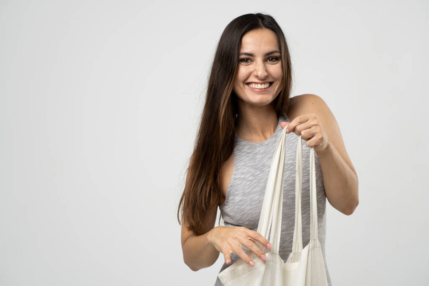 Cheerful millennial woman showing white eco bag to camera standing over white studio background. Lady holding cotton shopper handbag. fashion and ecology concept. - Foto, afbeelding