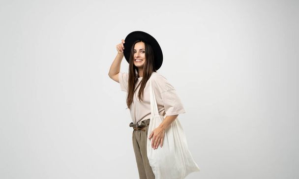 Brunette woman in a beige t-shirt and black hat with a cotton shopper bag with groceries looking in a camera. Reusable eco bag for shopping. Zero waste concept. - Foto, Imagen