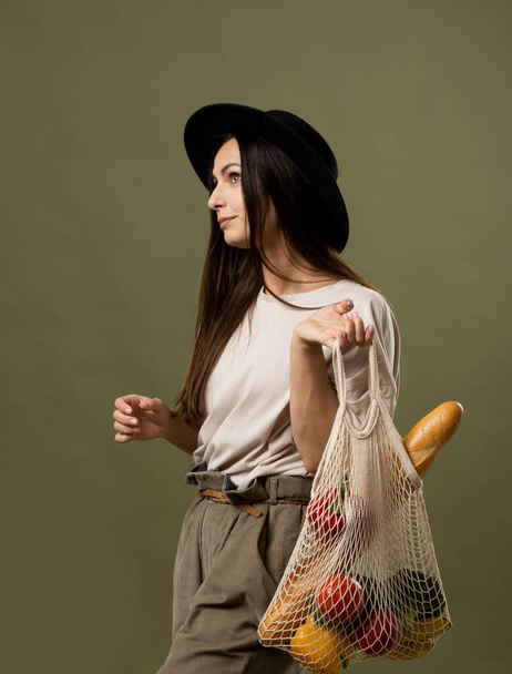 Eco friendly shopping. Smiling young woman holding string reusable mesh bag with organic fruits and vegetables. Positive female standing on green studio background. - Φωτογραφία, εικόνα