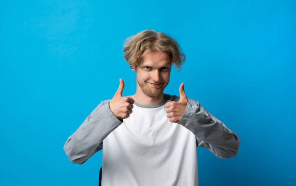 Young handsome blonde man in white t-shirt doing happy thumbs up gesture with both hand. Approving expression looking at the camera, showing success. - Photo, Image