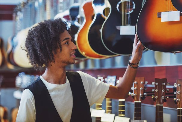 Bello giovane afro-americano che sceglie la chitarra e sorride in piedi in un negozio di musica
 - Foto, immagini