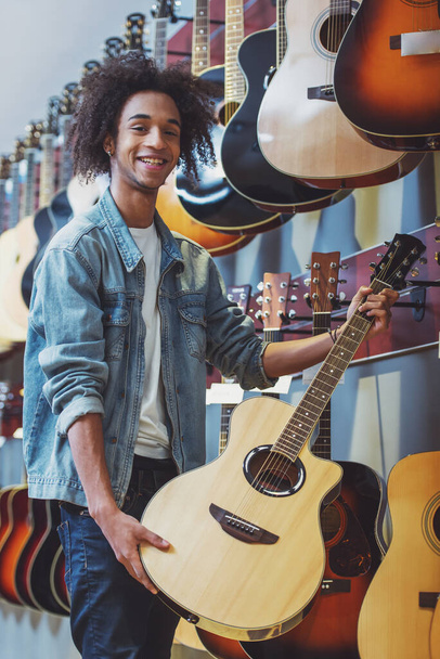 Όμορφος Αφροαμερικανός άντρας στο τζιν σακάκι κρατώντας μια κιθάρα, κοιτάζοντας την κάμερα και χαμογελώντας ενώ στέκεται σε ένα μουσικό κατάστημα - Φωτογραφία, εικόνα