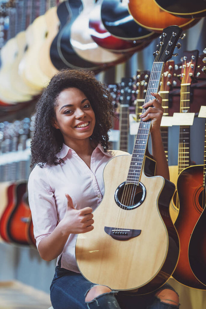 Szép afro-amerikai lány gazdaság egy gitár, mutatja az Ok jele, és mosolyogva, miközben ül egy zenei bolt - Fotó, kép