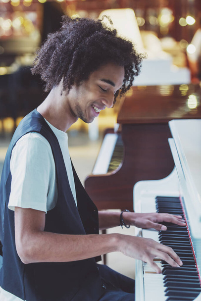 Vista lateral del guapo afroamericano en chaleco clásico sonriendo y tocando un piano en una tienda musical
 - Foto, imagen
