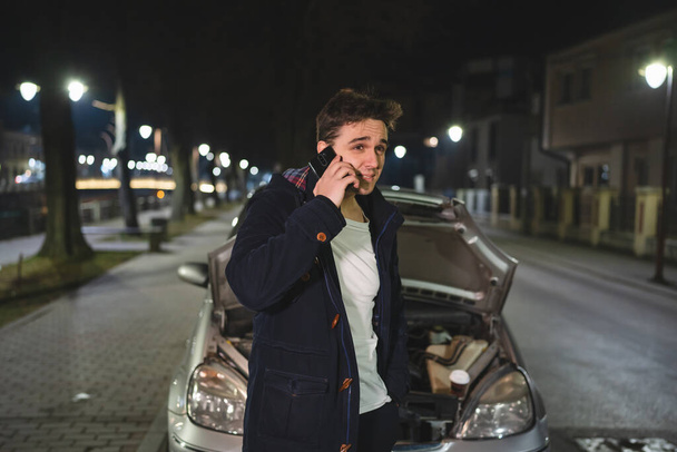 Um jovem homem caucasiano adulto em pé junto ao seu veículo com capuz aberto e motor avariado falhou segurando um telefone chamando serviço de reboque para ajuda na estrada Conceito de assistência Roadside na noite escura - Foto, Imagem