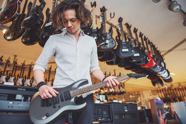 Knappe stijlvolle man spelen een elektrische gitaar terwijl je in een muzikale winkel - Foto, afbeelding