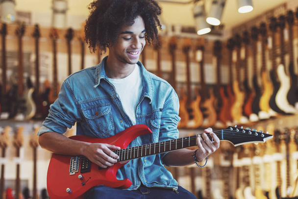 schöner junger afroamerikanischer Mann in Jeansjacke lächelt, während er in einem Musikladen eine E-Gitarre spielt - Foto, Bild