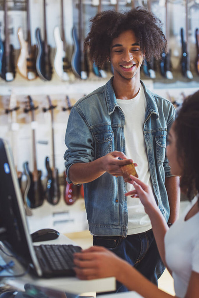 schöner junger afroamerikanischer Mann in Jeansjacke lächelt beim Bezahlen mit Kreditkarte in einem Musikladen - Foto, Bild