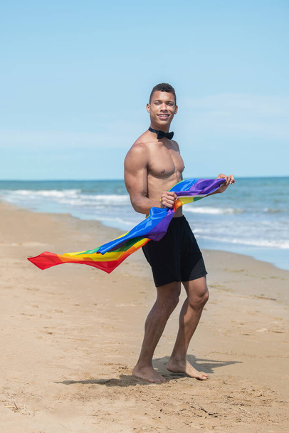 Man holding a rainbow gay pride flag while standing on the beach. - Фото, зображення