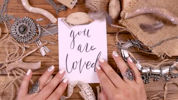 Widok z góry młodej kobiety dotykającej odręcznie napisaną nutę słowami You Are LOVED na drewnianym stole z etniczną biżuterią i naturalnym wystrojem. Ręcznie robione akcesoria rzemieślnicze, miłość, Valentine 's concept - Materiał filmowy, wideo