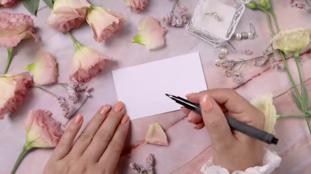 Mains qui écrivent BRIDE sur une carte près de fleurs roses près d'une table en marbre - Séquence, vidéo