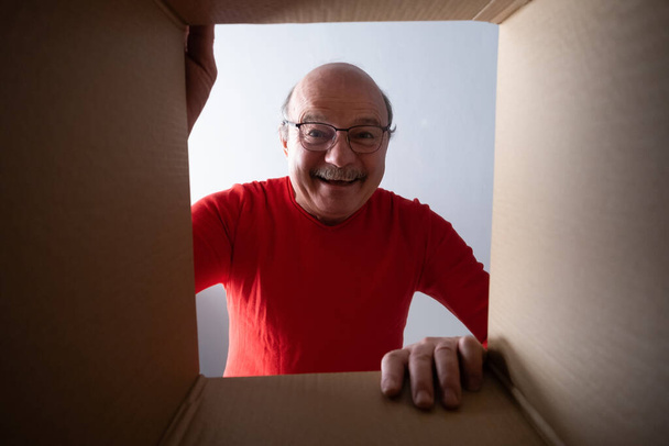Starší překvapený muž vybaluje, otevírá kartónovou krabici a nahlíží dovnitř. - Fotografie, Obrázek