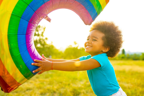 positive multicultural race little girl with afro curly hair holding rainbow balloon in summer park - 写真・画像