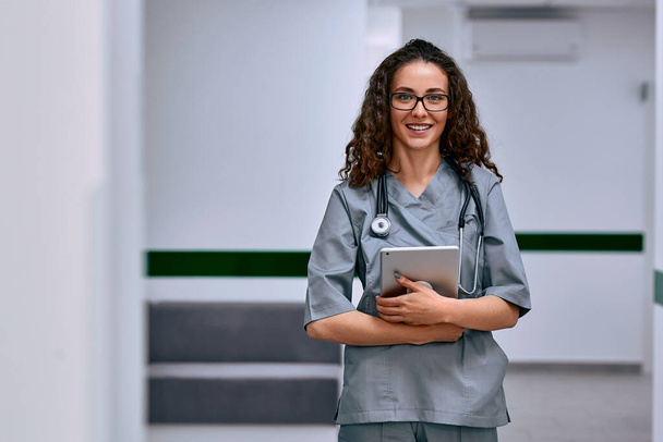 A female doctor is wearing a medical suit and glasses, a stethoscope around her neck, holding a tablet and standing in the hospital corridor, looking at the camera and smiling.Copy space. - Fotó, kép
