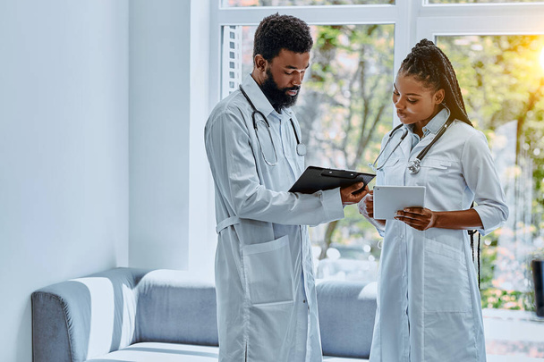 Deux jeunes médecins en blouse blanche discutent du traitement dans le hall de l'hôpital et regardent la tablette. - Photo, image