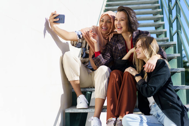 Happy multiethnic female best friends taking selfie sitting on stairs in - Photo, Image