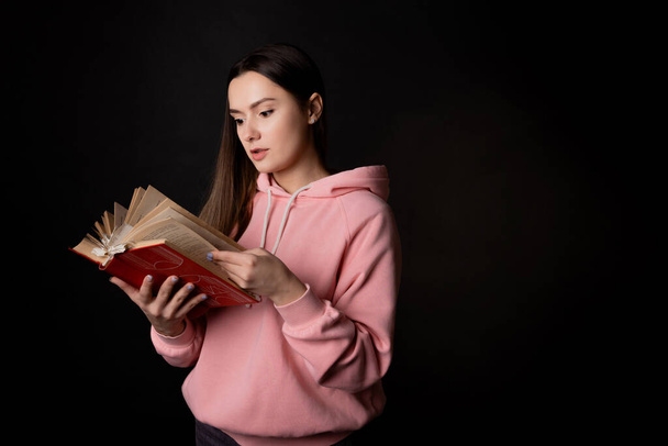 Een jonge, schattige brunette met een roze capuchon, die een boek leest. Studente met een open boek in haar handen, - Foto, afbeelding