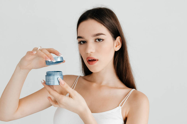 Beautiful brunette woman with perfect nude makeup, wearing white top, posing with a blue cream jar on white background. Beauty concept - Foto, Bild