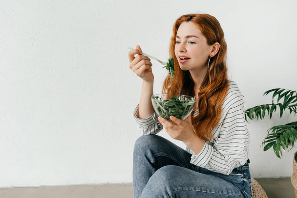 Lekker en gezond maaltijdconcept. Een roodharige vrouw in casual kleding eet een verse salade uit een kom - Foto, afbeelding