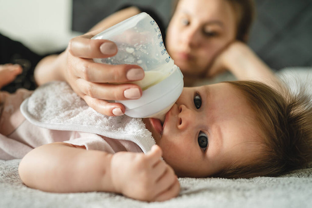 Κοντινό πλάνο σε μικρό καυκάσιο μωρό τεσσάρων μηνών ξαπλωμένο στο κρεβάτι στο σπίτι, ενώ η μητέρα της την ταΐζει με μπουκάλι γάλα φόρμουλα μητρότητα και την έννοια των γονέων πραγματικούς ανθρώπους - Φωτογραφία, εικόνα