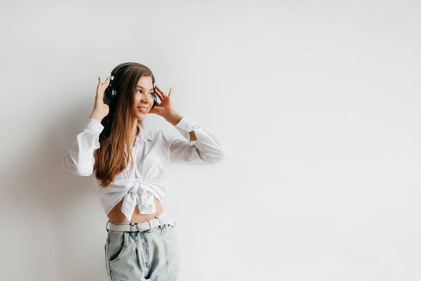 Beautiful young brunette woman in white shirt and jeans posing over white background. Teenage lifestyle concept. Listening to music with headphones - Valokuva, kuva