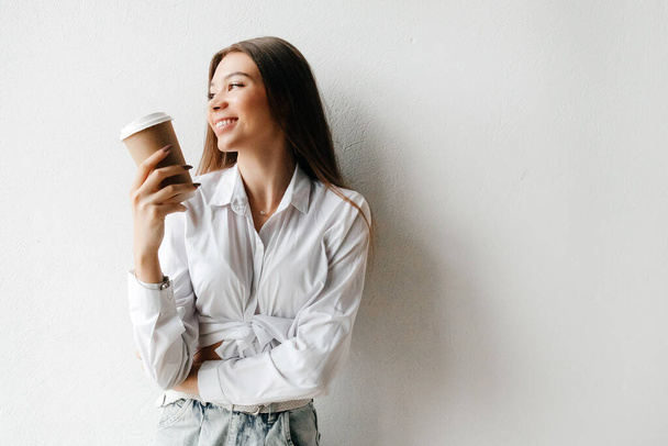 Młoda brunetka kobieta trzyma jednorazowy kubek kawy i uśmiecha się, patrząc z boku - Zdjęcie, obraz