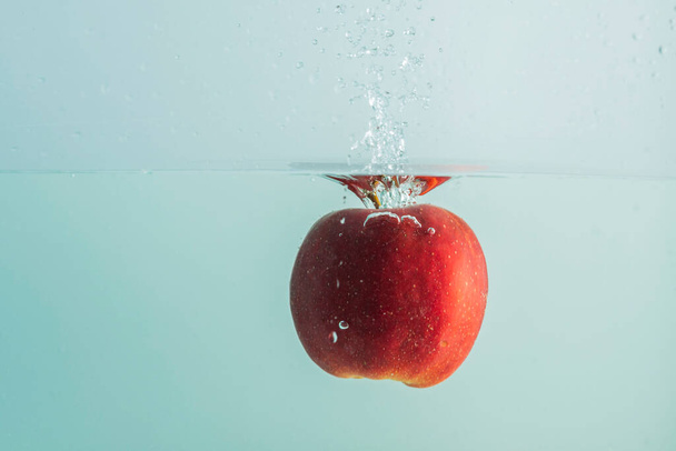Закройте вид красного яблока, падающего в воду на бирюзовом фоне. Великолепный фон. - Фото, изображение