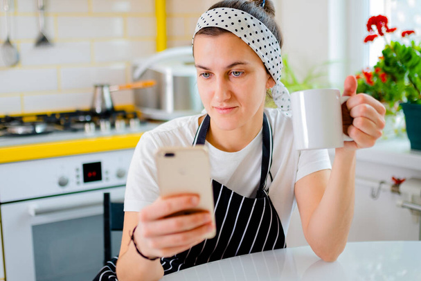kobieta siedzi przy stole i używa smartfona do oglądania wiadomości, rozmawiać podczas relaksu w kuchni. koncepcja relaksu - Zdjęcie, obraz