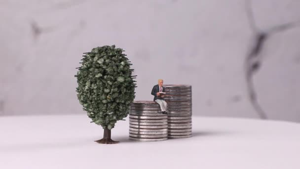 Un uomo in miniatura che legge un libro su un mucchio di monete e un albero in miniatura. Miniature e pile di monete con concetti di business. - Filmati, video
