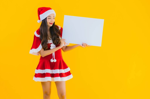 Güzel genç Asyalı kadın portresi Noel kıyafetleri ve sarı izole edilmiş arka planda boş beyaz pano işareti olan şapka gülüşü. - Fotoğraf, Görsel