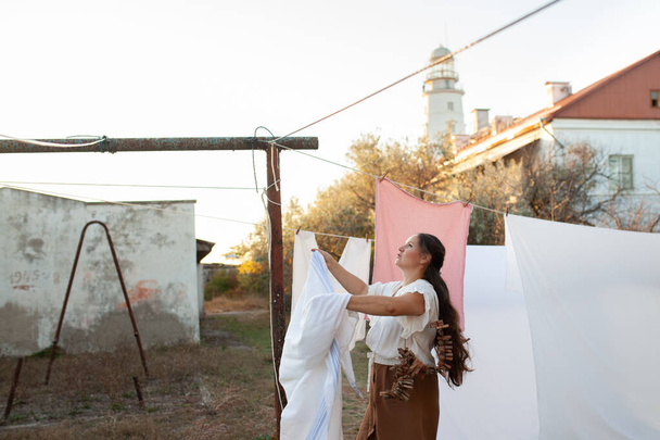 beautiful woman with long hair hangs white sheets in the air near the lighthouse, on the street a warm autumn day, sunset, evening, outdoor - Φωτογραφία, εικόνα