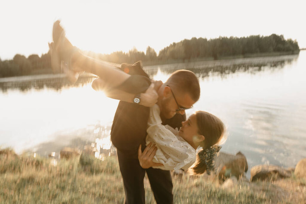A blurred photo of a young father with a beard who is circling his daughter in his arms on the coast of the lake. Single-parent family on vacation on the sunset. - Photo, Image