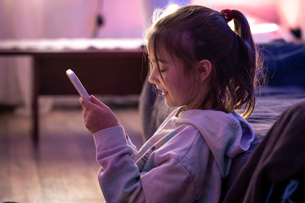 Ένα μικρό κορίτσι χρησιμοποιεί ένα smartphone, ενώ κάθεται στο δωμάτιό της. - Φωτογραφία, εικόνα