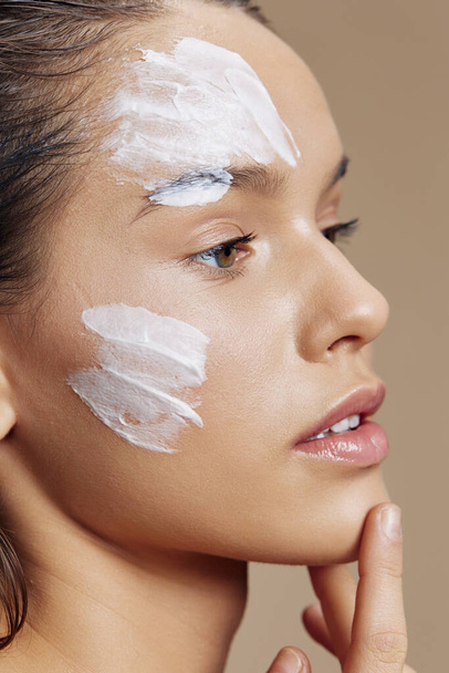 young woman with moisturizer on the cheeks in the eye area beige background - Zdjęcie, obraz