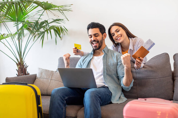 Χαρούμενοι νεαροί τουρίστες κάνουν κράτηση δωματίου ξενοδοχείου online, χρησιμοποιώντας φορητό υπολογιστή, γυναίκα που κατέχει πιστωτική κάρτα και διαβατήρια με εισιτήρια - Φωτογραφία, εικόνα