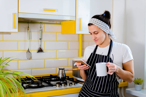 Kobieta używająca smartfona i pijąca kawę w kuchni. Concept szczęśliwy poranny wypoczynek - Zdjęcie, obraz