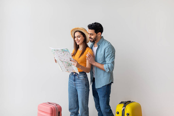 Happy travelers couple planning vacation and using tourist map, choosing destination for trip, standing over light wall - Photo, Image