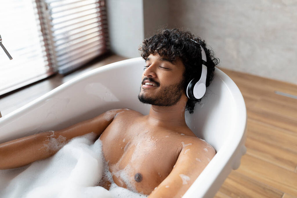 Entspannter indischer Mann hört beruhigende Musik in drahtlosen Kopfhörern und liegt mit geschlossenen Augen im Schaumbad - Foto, Bild