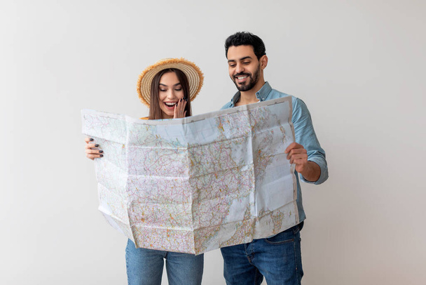 Jonge echtgenoten die reisbestemming kiezen voor vakantie, staan met toeristische kaart over lichtmuur, vrije ruimte - Foto, afbeelding