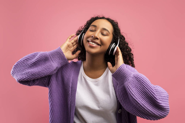 ピンクのスタジオの背景にヘッドフォンを身に着けて、閉じた目で音楽を聴く楽しい千年の黒人女性 - 写真・画像