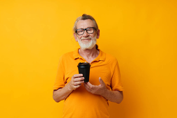 Φωτογραφία του συνταξιούχου γέρου με ένα κίτρινο T-shirt ένα ποτήρι με ένα ποτό απομονωμένο φόντο - Φωτογραφία, εικόνα