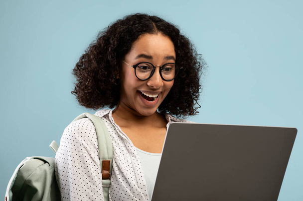 Συναισθηματική νεαρή Αφροαμερικανή γυναίκα με σακίδιο κοιτάζοντας οθόνη laptop σε ενθουσιασμό πάνω από το μπλε φόντο - Φωτογραφία, εικόνα