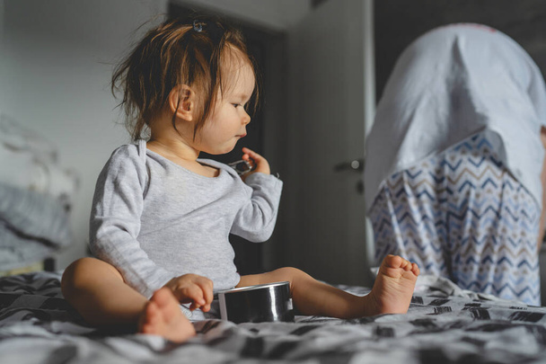 Yksi valkoihoinen tyttö yhdeksän kuukauden ikäinen leikkii sängyssä kotona päivällä tai aamulla äitinsä kanssa taustalla valitsemalla vaatteita kopioi tilaa lapsuus ja kasvaa perhekonsepti. - Valokuva, kuva