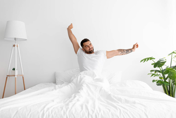 Tevreden jonge knappe man met baard ontwaakt en stretching lichaam op wit bed in slaapkamer interieur - Foto, afbeelding