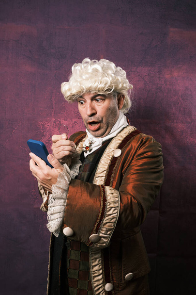 Homem em roupas renascentistas clássicas com uma expressão surpresa em seu rosto enquanto olha para a tela do telefone celular com um monóculo. - Foto, Imagem