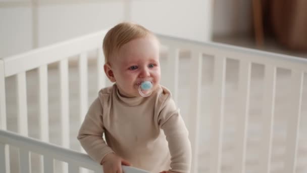 Очаровательный ребенок с успокаивающим средством во рту, стоящий в кроватке и машущий рукой - Кадры, видео