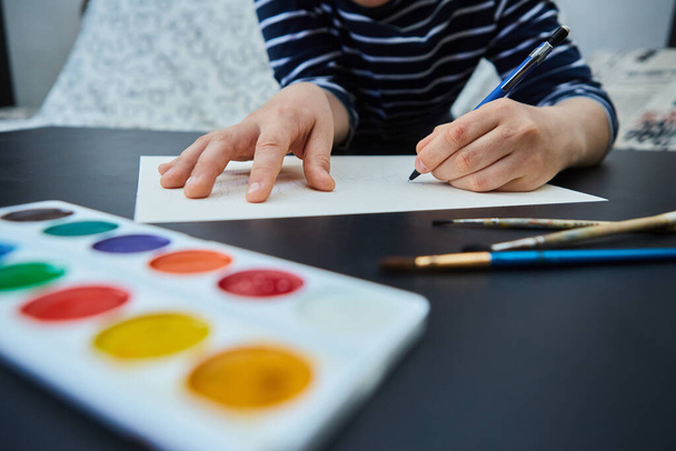 Las manos infantiles con el lápiz azul dibujan el dibujo sobre el papel blanco sobre la mesa de madera. Vista lateral. - Foto, imagen