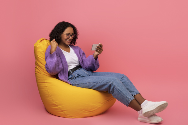 Emocionado joven mujer negra sentada en la silla de la bolsa de frijol con el teléfono inteligente, gesto SI, ganar apuesta de casino o juego en línea - Foto, Imagen